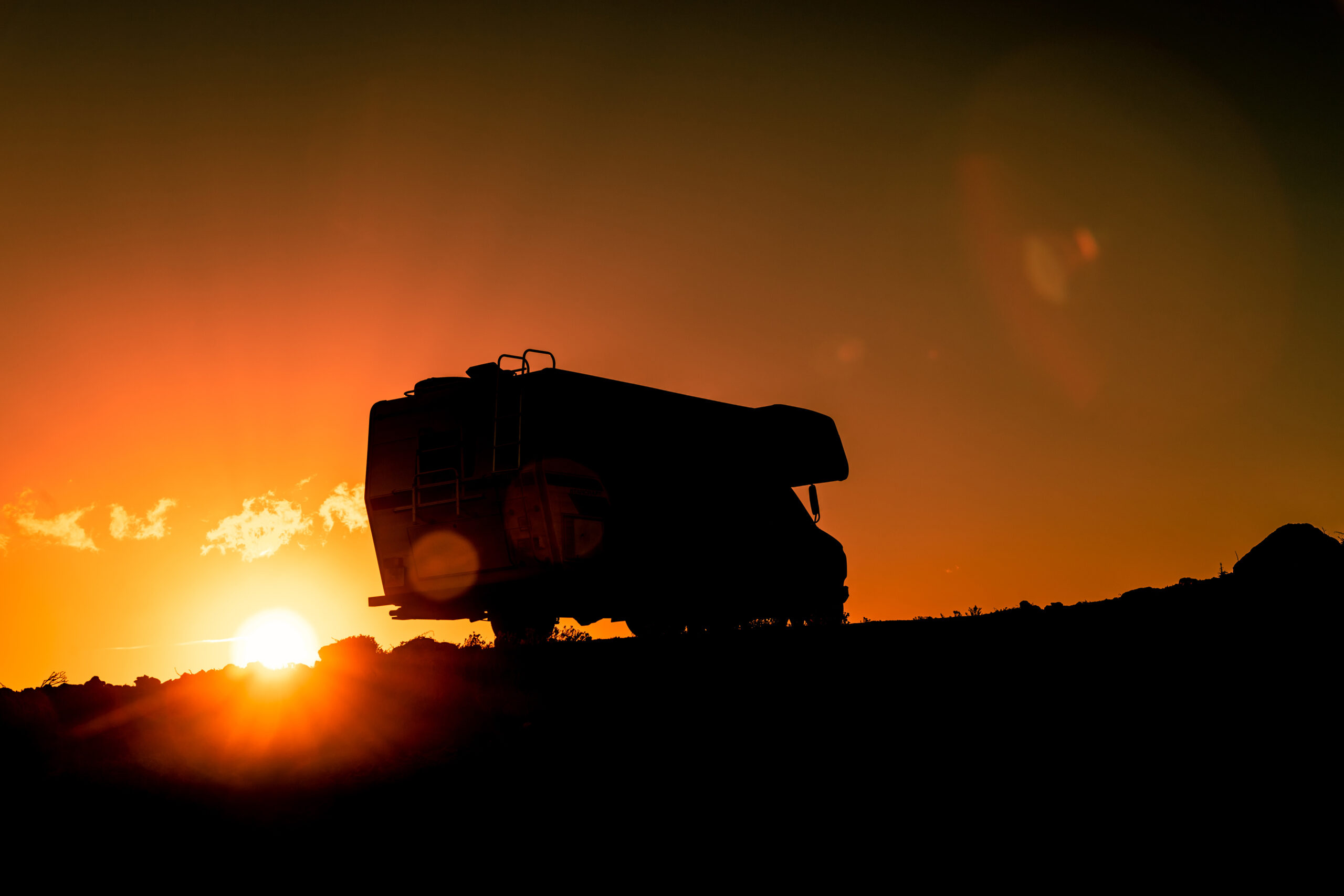 Silhouette of a truck on a hill at sunset, with the sun partially visible on the horizon and the sky glowing orange. The truck appears large and is positioned on the right side of the image.