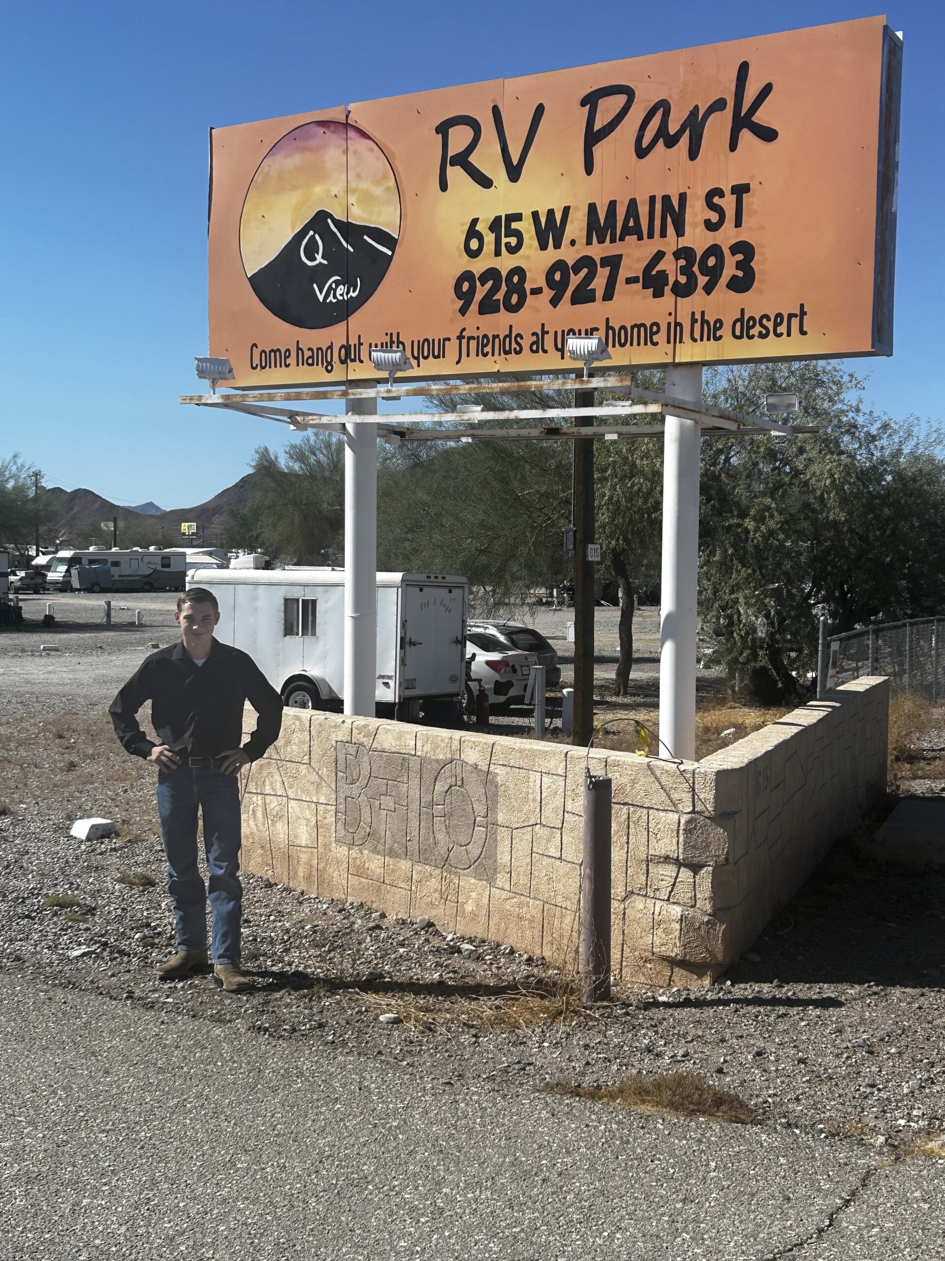Man standing near a roadside sign for an RV park that reads, "RV Park, 615 W. Main St, 928-927-6393, Come hang out with your friends at your home in the desert." Desert landscape and a trailer are visible in the background.