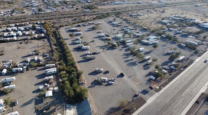 Aerial view of an RV campground with numerous RVs parked on a dirt lot. Sparse trees and desert vegetation are visible. Roads border the campground, with a railway track in the background, and a few buildings scattered throughout.