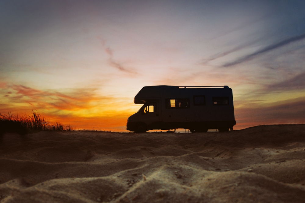 Silhouette of a camper van parked on sandy terrain during sunset. The sky is painted with vibrant hues of orange, pink, and purple. Grasses are visible on the horizon, adding to the serene landscape.